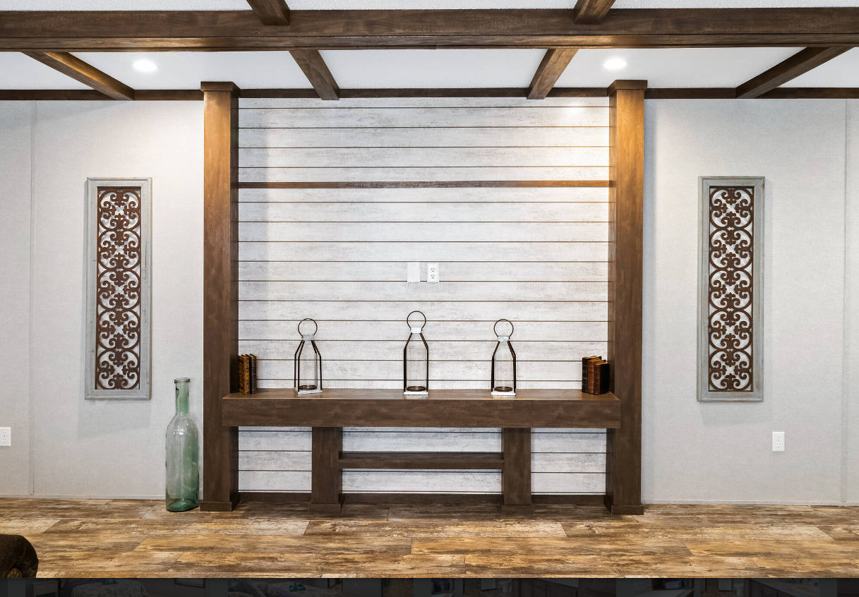 Decorative wall with three lanterns, two books, and a tall glass vase on a wooden shelf against a striped white wood panel background, flanked by two ornate metal wall art pieces on light-colored walls.