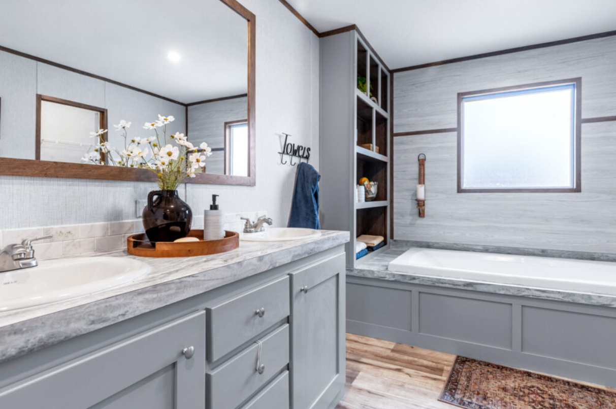 Bathroom with light gray cabinetry, marble countertops, a sink, a mirror, a vase with white flowers, wooden tray, soap dispenser, towel on a hook, open shelves with decor, frosted window, and a bathtub with a wood-accented wall.