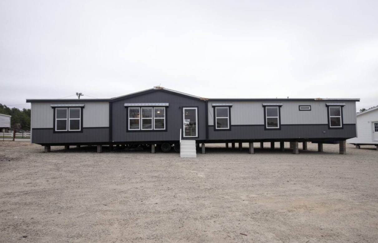 A modern manufactured home elevated on stilts, painted in dark and light gray, with steps leading up to the front door and multiple windows.