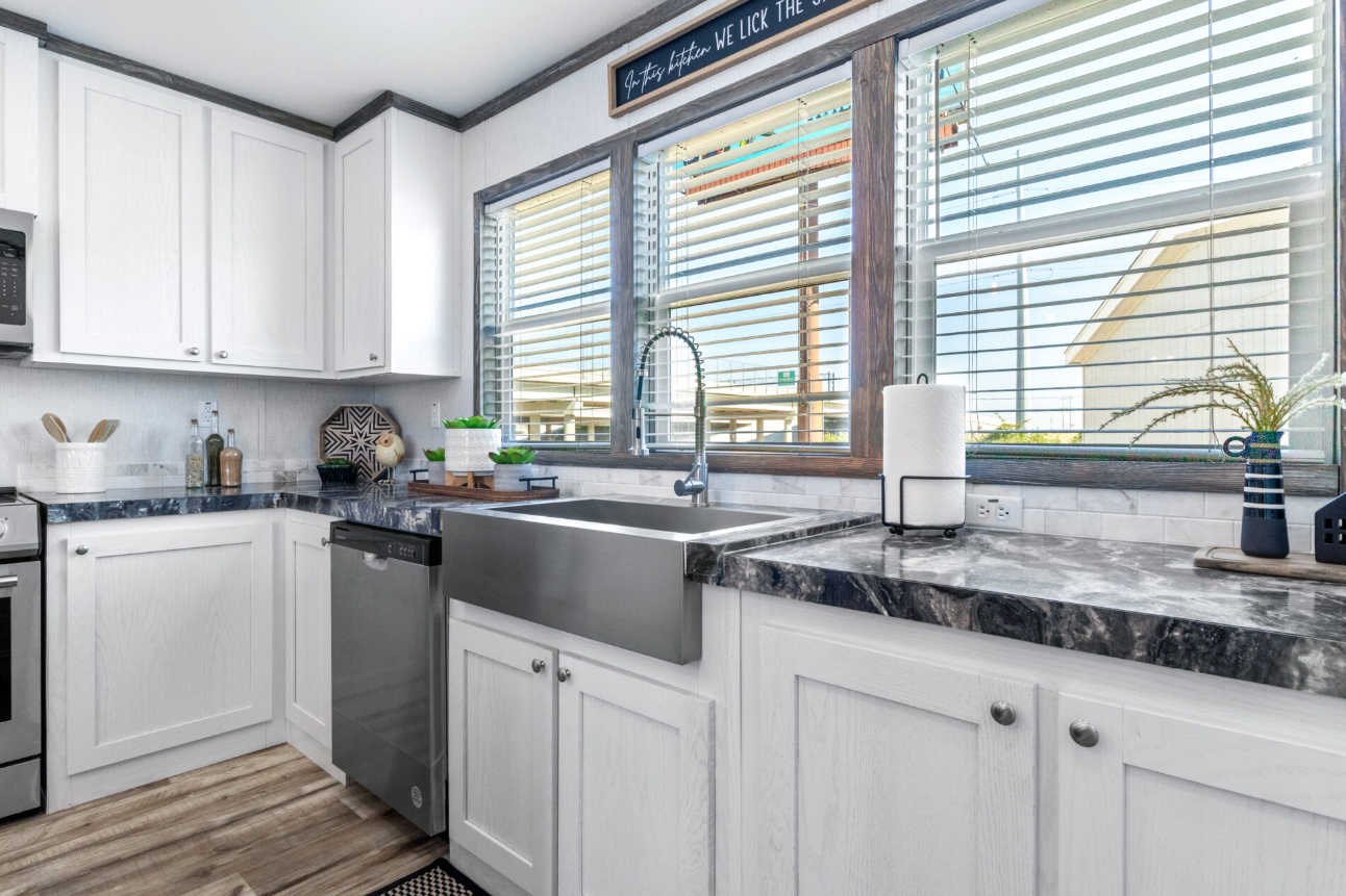 Modern kitchen with white cabinets, black marble countertops, a farmhouse-style stainless steel sink, and large windows with wooden trim bringing in natural light. Countertop decor includes small potted plants, a paper towel holder, and a vase with d