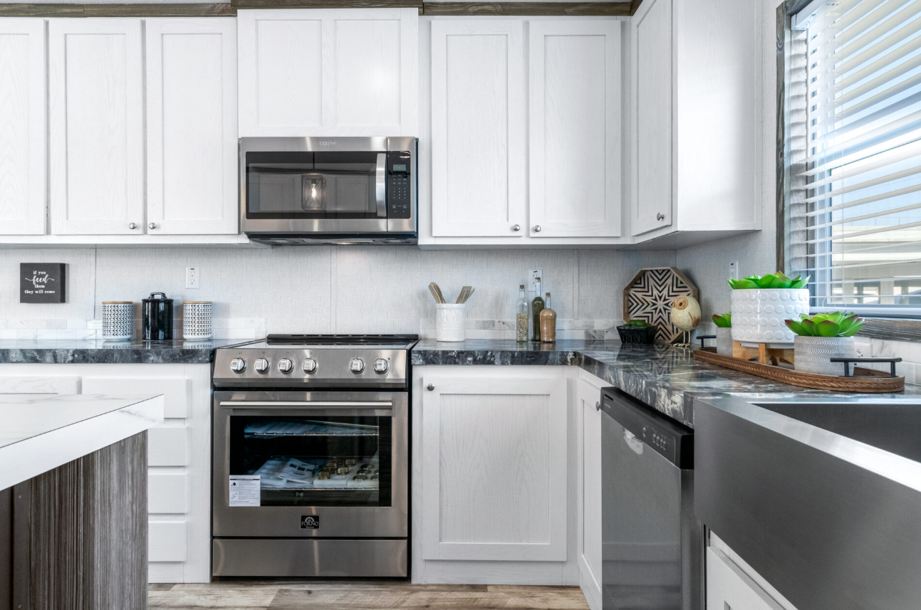 Modern kitchen with white cabinets, black marble countertops, stainless steel appliances including a microwave, oven, and dishwasher, with a window to the right decorated with green plants.