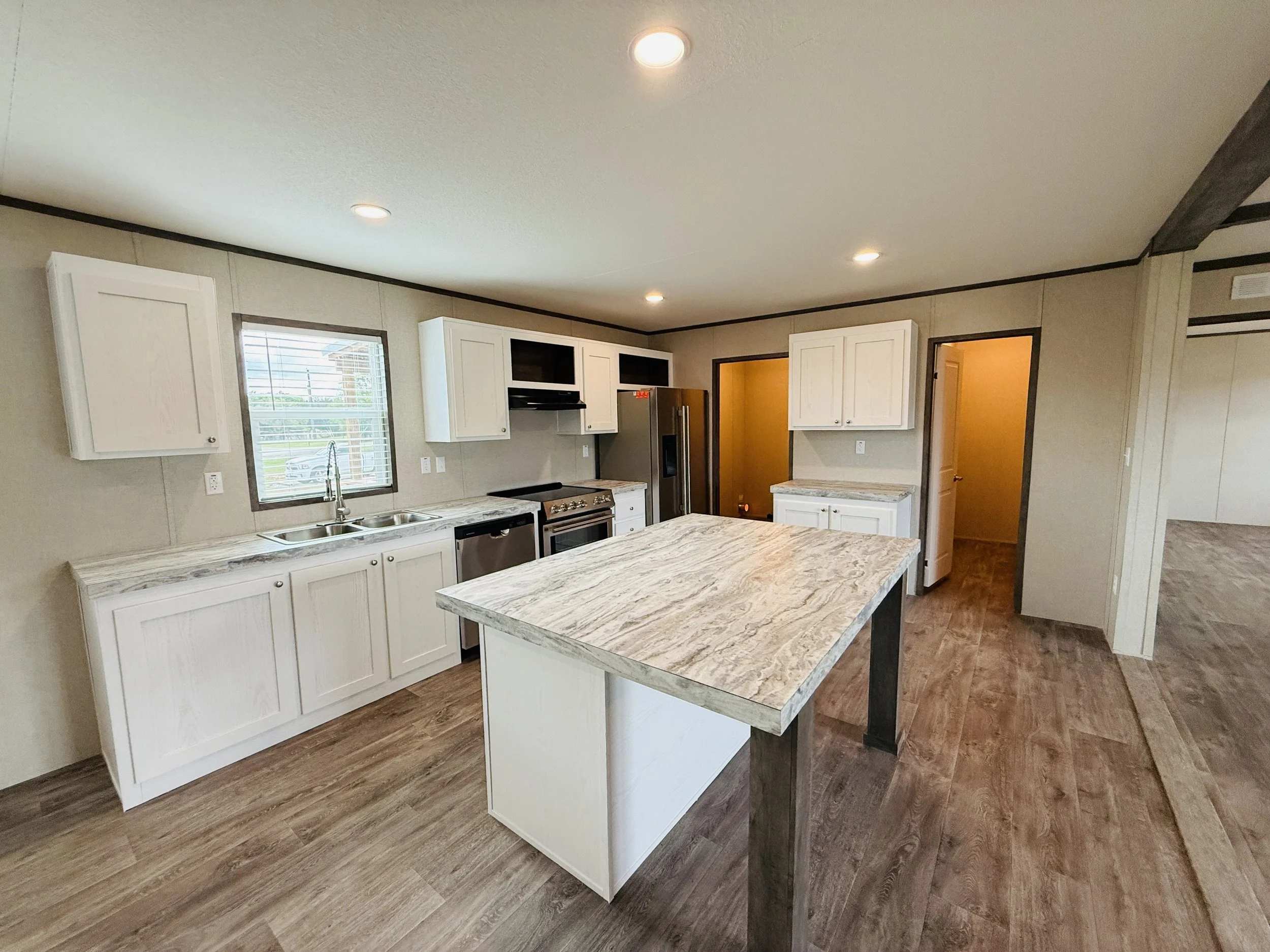 Modern kitchen with white cabinets, marble countertops, and an island, stainless steel appliances, wooden flooring, and a sunlit window.