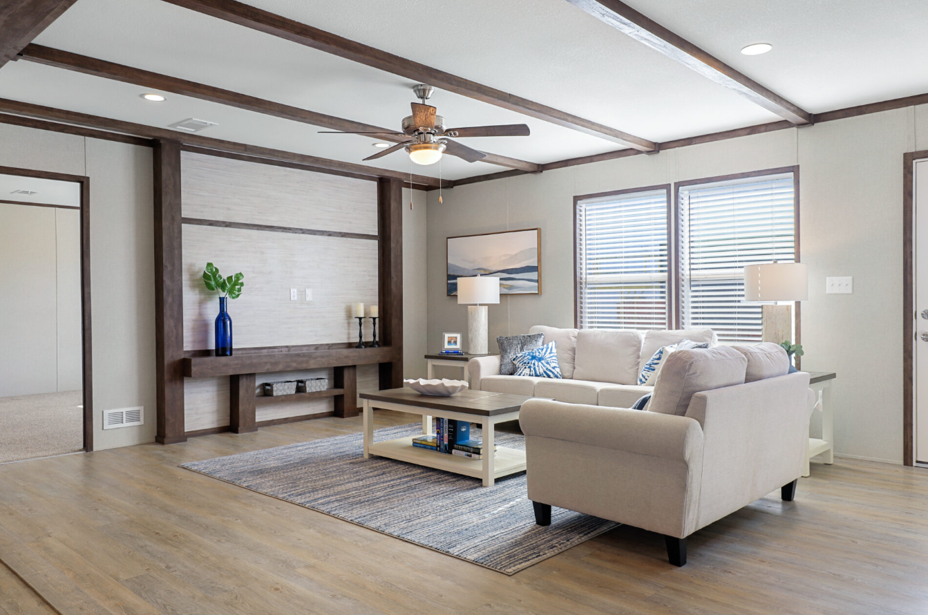 A living room with beige sofas, a wooden coffee table, a corner bookshelf, and two side tables with lamps, all illuminated by natural light from two windows and decorated with blue accents and wall art.