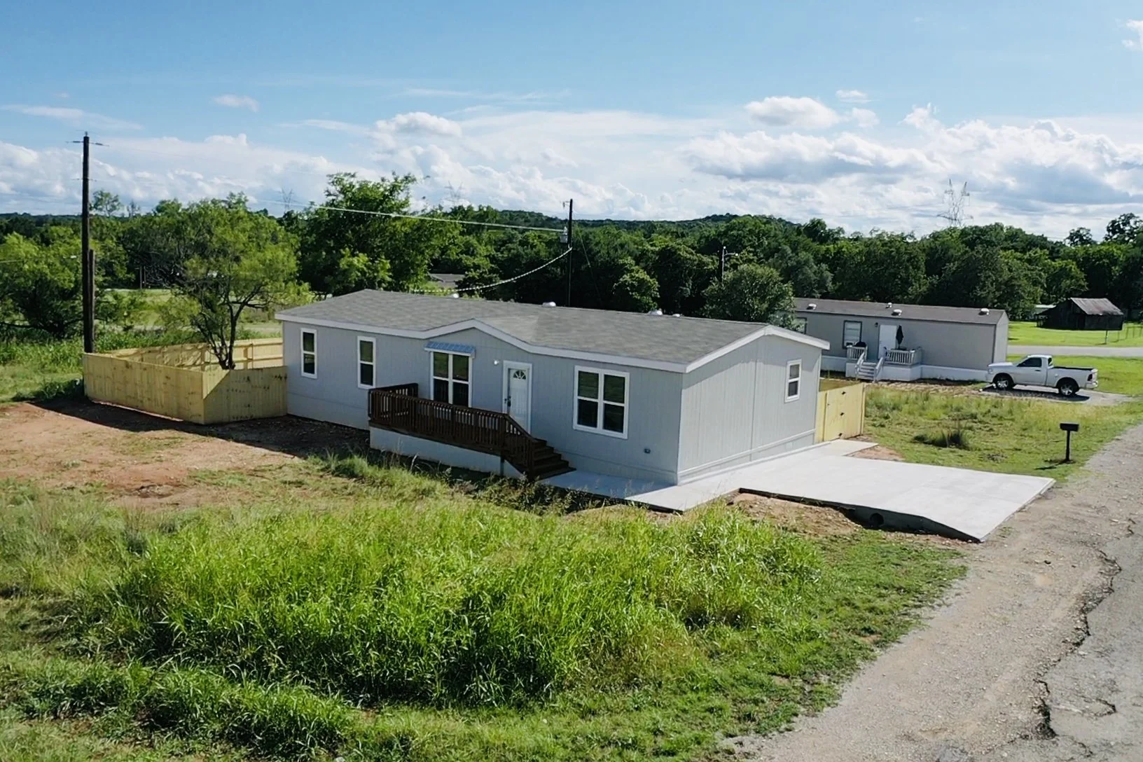 Newly built single-story mobile home with a gray exterior, two front windows, a small front deck with stairs, situated on a lot with a concrete driveway, surrounded by grass, with another mobile home and a pickup truck in the background, under a part