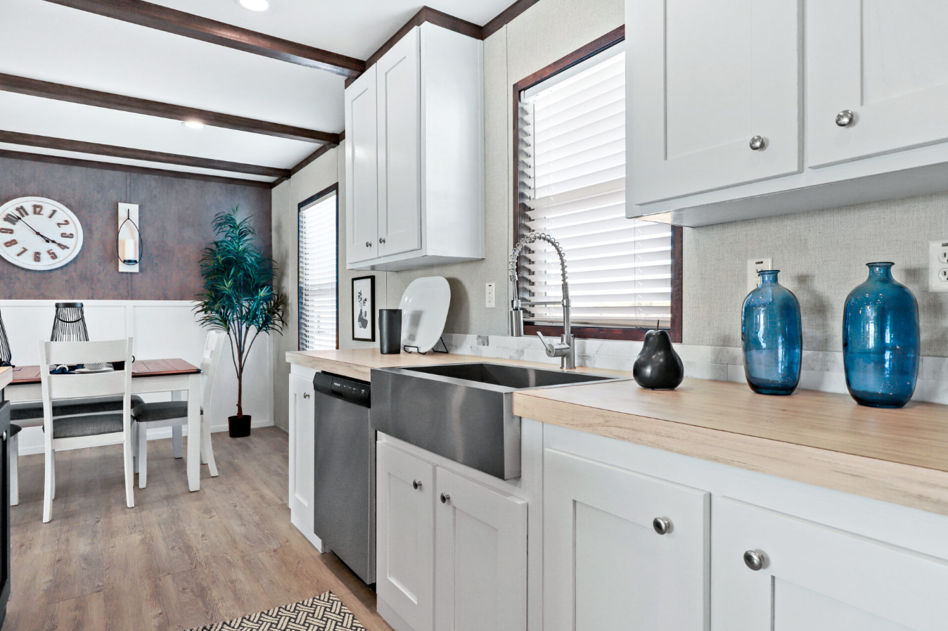 Kitchen with white cabinets, farmhouse sink, blue vases, patterned rug, dining table, clock, and potted plant.