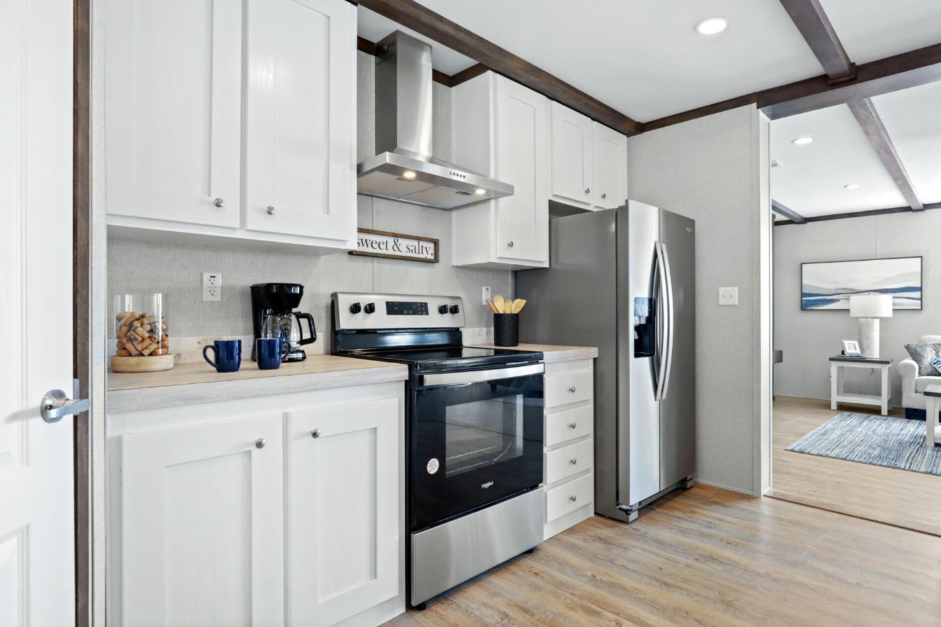 Modern kitchen with white cabinets, stainless steel refrigerator and stove, black coffee maker, two blue mugs, a jar of cookies, a utensil holder, and a decorative sign that says 'sweet & salty'.