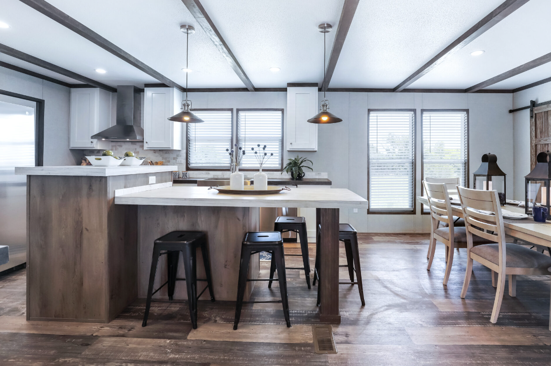 Open-concept kitchen and dining area with a central wooden kitchen island, black bar stools, white cabinetry, stainless steel appliances, and a dining table with chairs near large windows.
