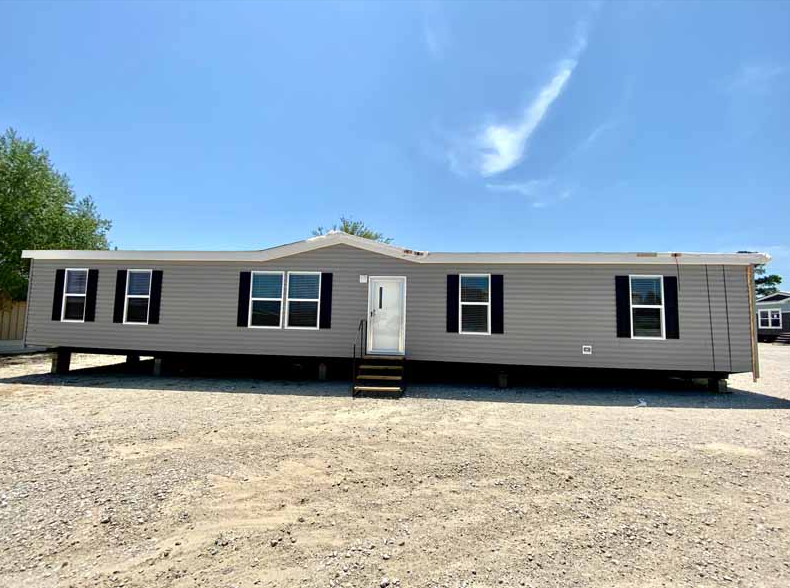 Single-story manufactured home with gray siding, black shutters, and a front door with steps, set on a gravel lot under a blue sky.