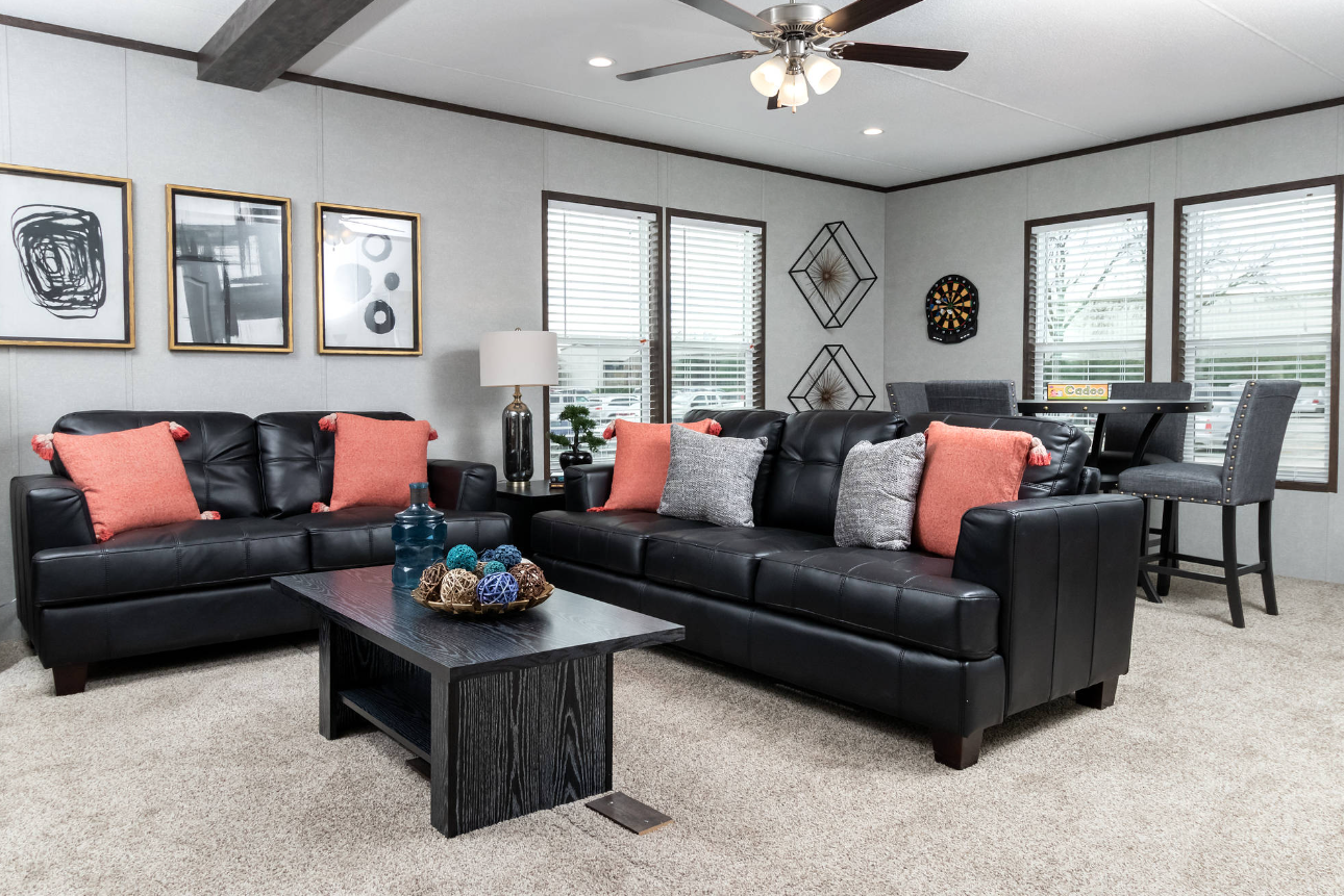 Living room with two black leather sofas and throw pillows, a black coffee table with decorative balls and vases, a side table with a lamp and plant, and a dining nook with a table and four chairs near multiple windows with blinds.