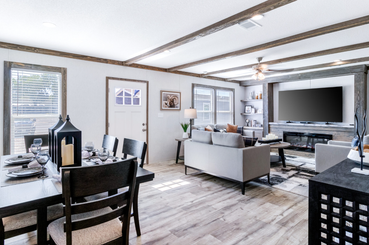 Open concept living room and dining area with white walls, wooden beams, a mounted flat-screen TV, beige sofas, black coffee table, and dining table set for four with black chairs and candle centerpiece