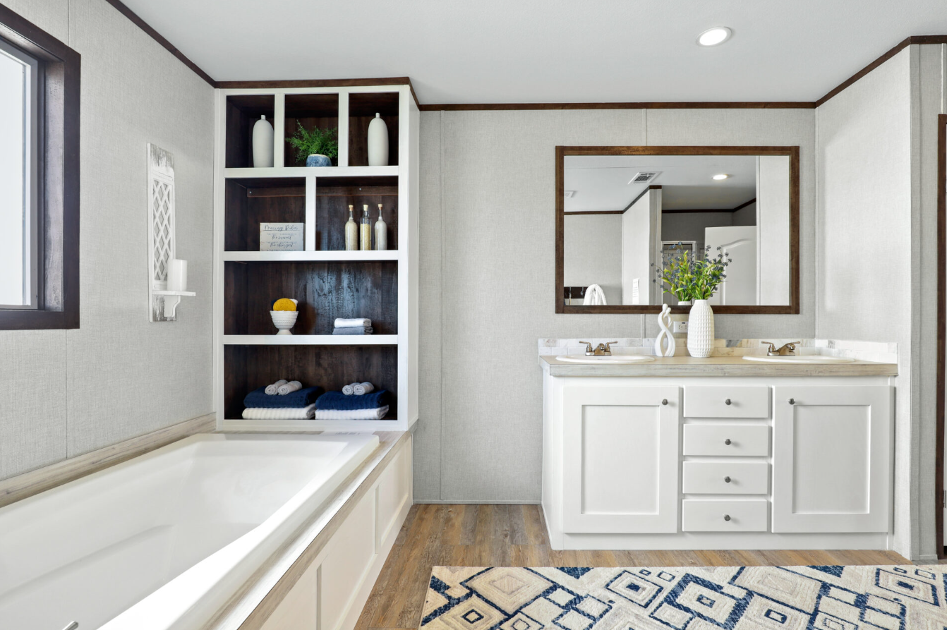 Bathroom with a large bathtub on the left, a white shelf with decorative items, and a double sink vanity with a large mirror on the right. There is a window next to the bathtub and a patterned rug on the wooden floor.