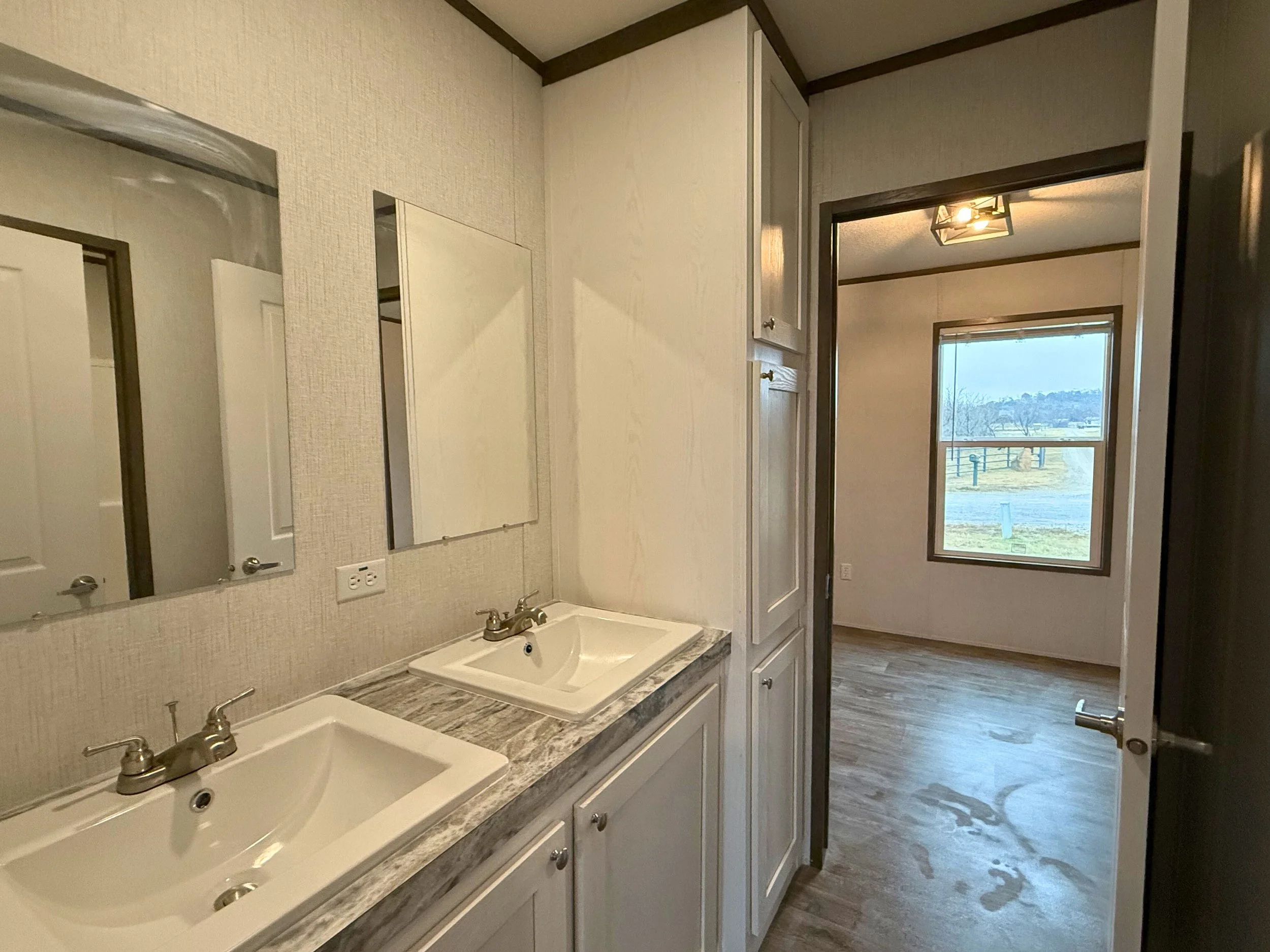 Bathroom with dual sinks, mirrors, and a doorway leading to a room with a large window showing an outdoor view.