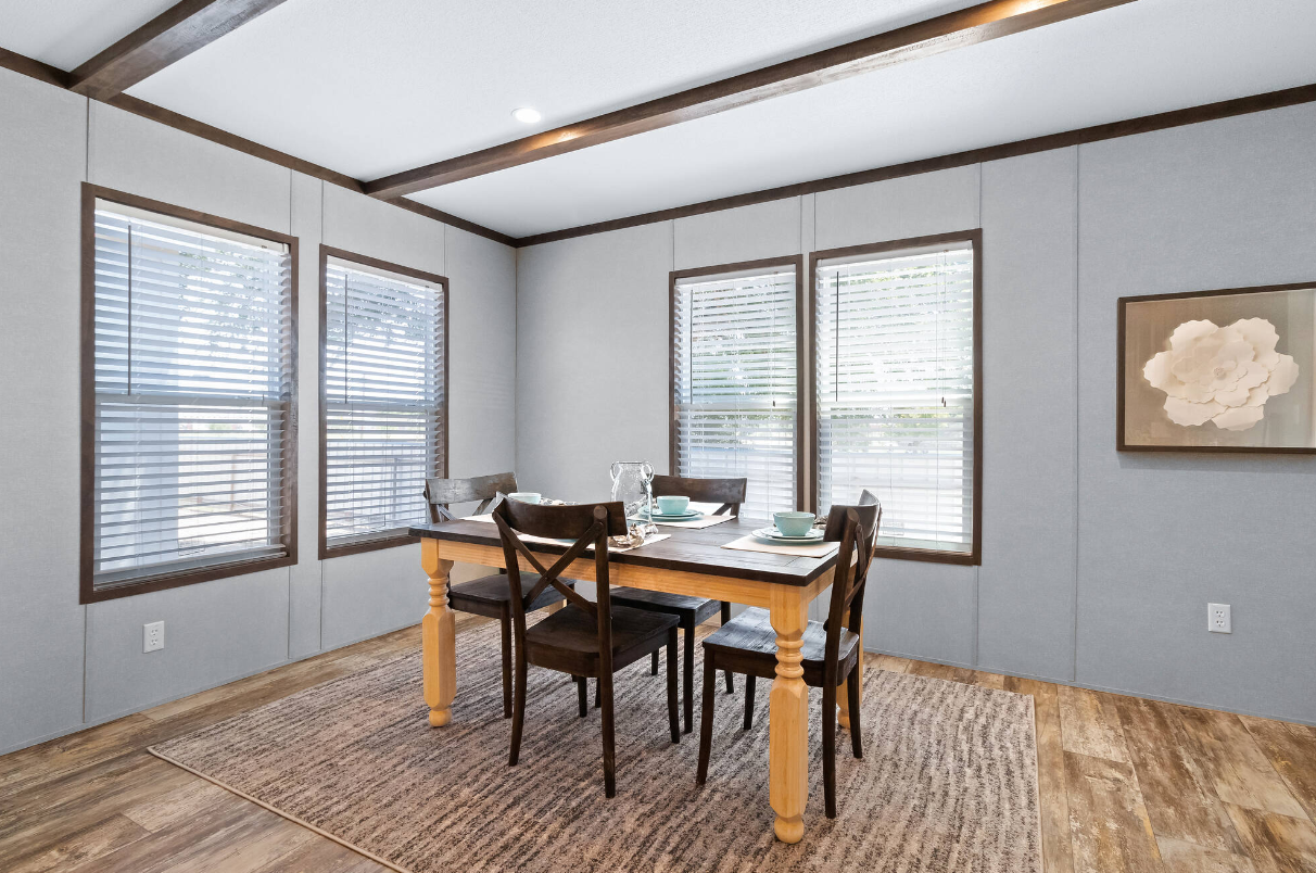 A dining room with a wooden table set for four, four chairs, three windows with blinds, a framed floral artwork on the wall, and hardwood flooring with an area rug.