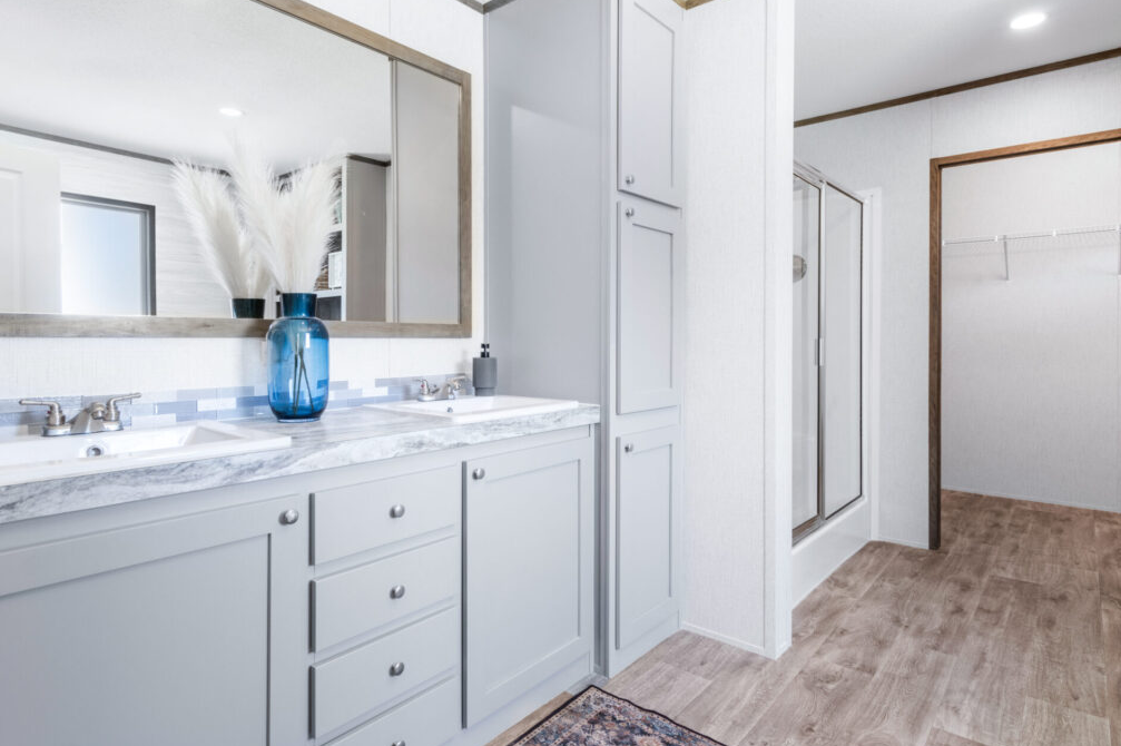 Bathroom with white cabinetry, large mirror, marble countertop, blue vase with white pampas grass, and glass shower enclosure, light-colored wood flooring, and white walls.