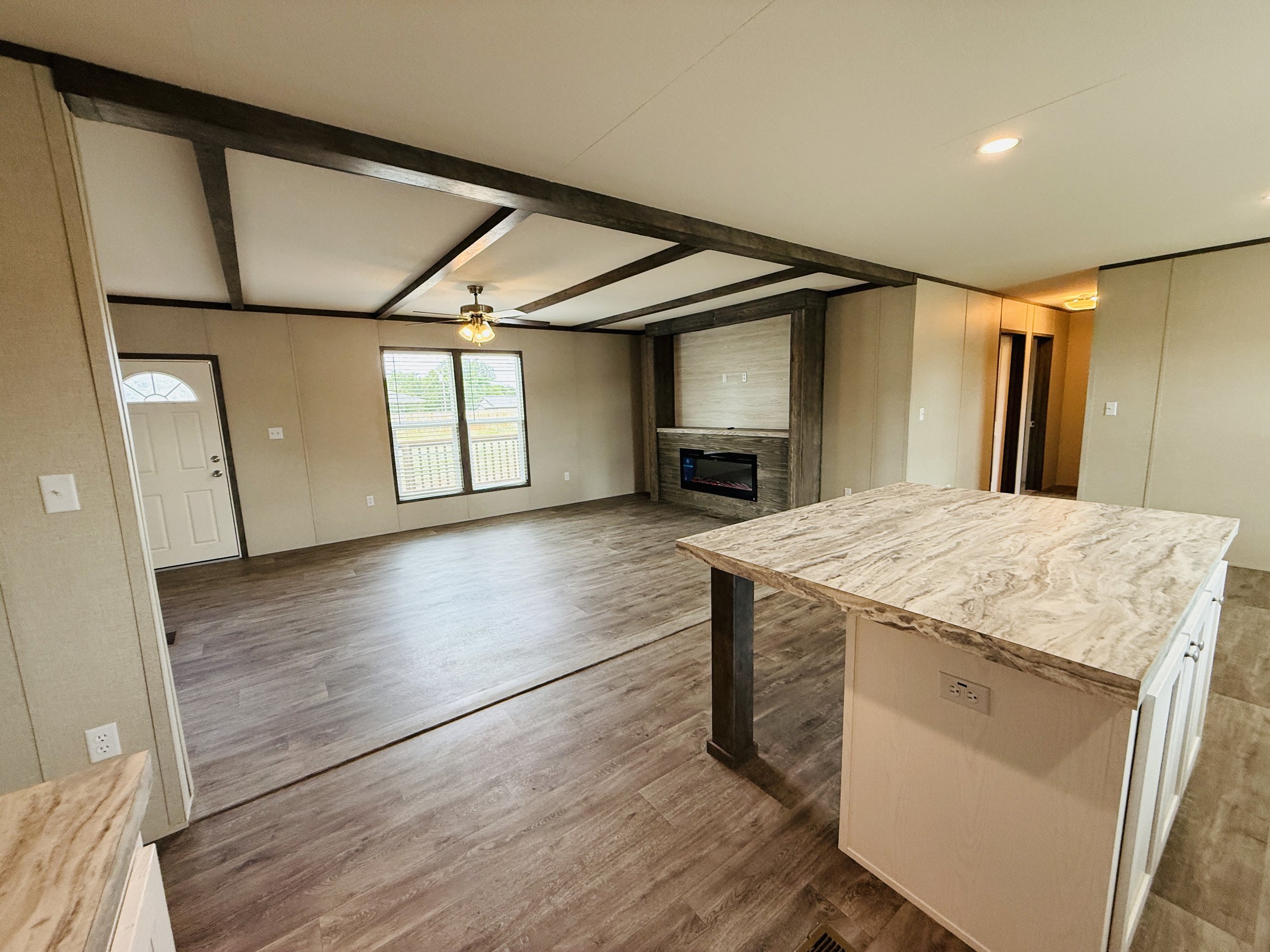 Empty living room with wood flooring, fireplace, large windows, beige walls, ceiling fan, and a kitchen island with marble countertop.