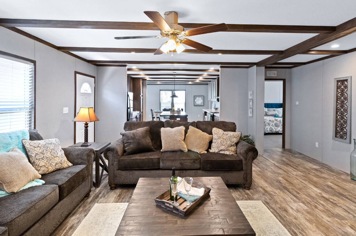 Living room with brown sofas, throw pillows, wooden coffee table with wine bottle, and a ceiling fan with lights. Open kitchen and dining area in the background.
