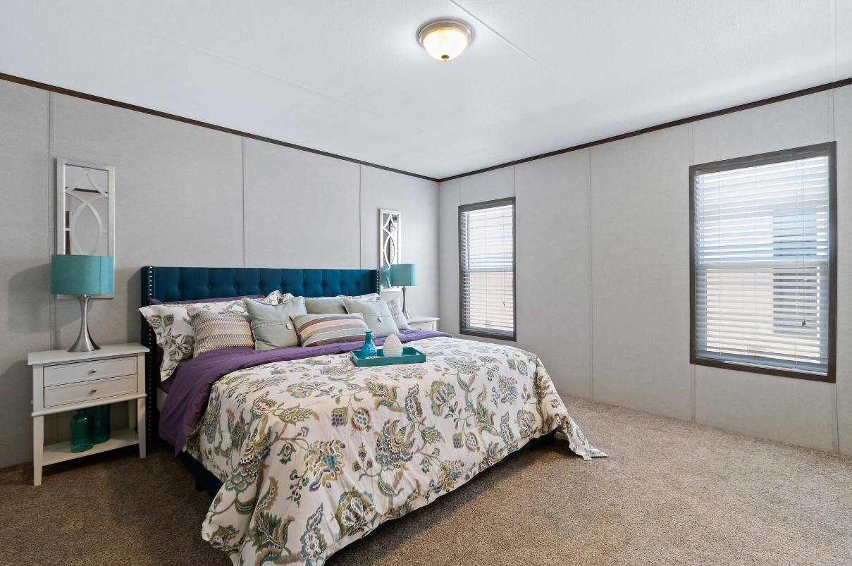 A bedroom with a bed that has a blue tufted headboard, decorative pillows, a floral quilt, and a tray with decorative items on it. There are two nightstands with teal lamps, and two windows with blinds. The room has beige carpet and gray walls.