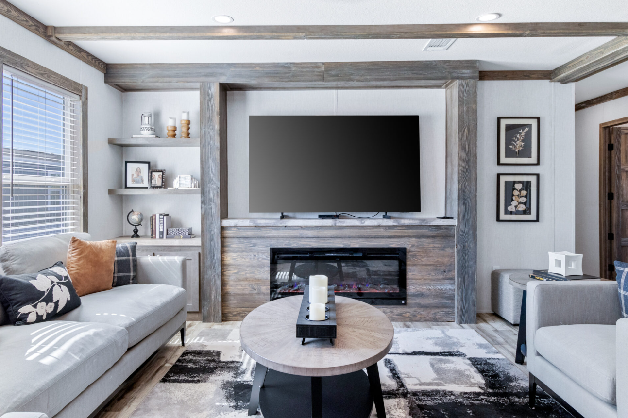 Living room with a wall-mounted TV above a modern fireplace, surrounded by light-colored walls and wood accents, with a white sofa on the left and a gray armchair on the right, and a round coffee table with candles in the center.