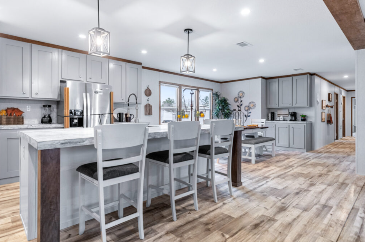 Modern kitchen with white cabinets, a marble island, and a wooden floor. There are four white chairs at the island, pendant lights hanging from the ceiling, and a window behind the sink with decorative plants.