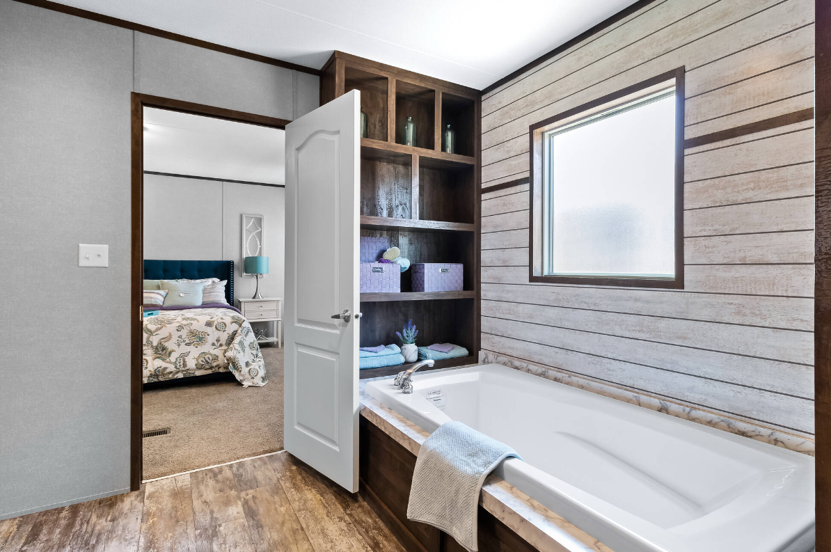Bathroom with bathtub, wood shelves with towels and decor, window, and doorway leading to bedroom with bed and nightstand.