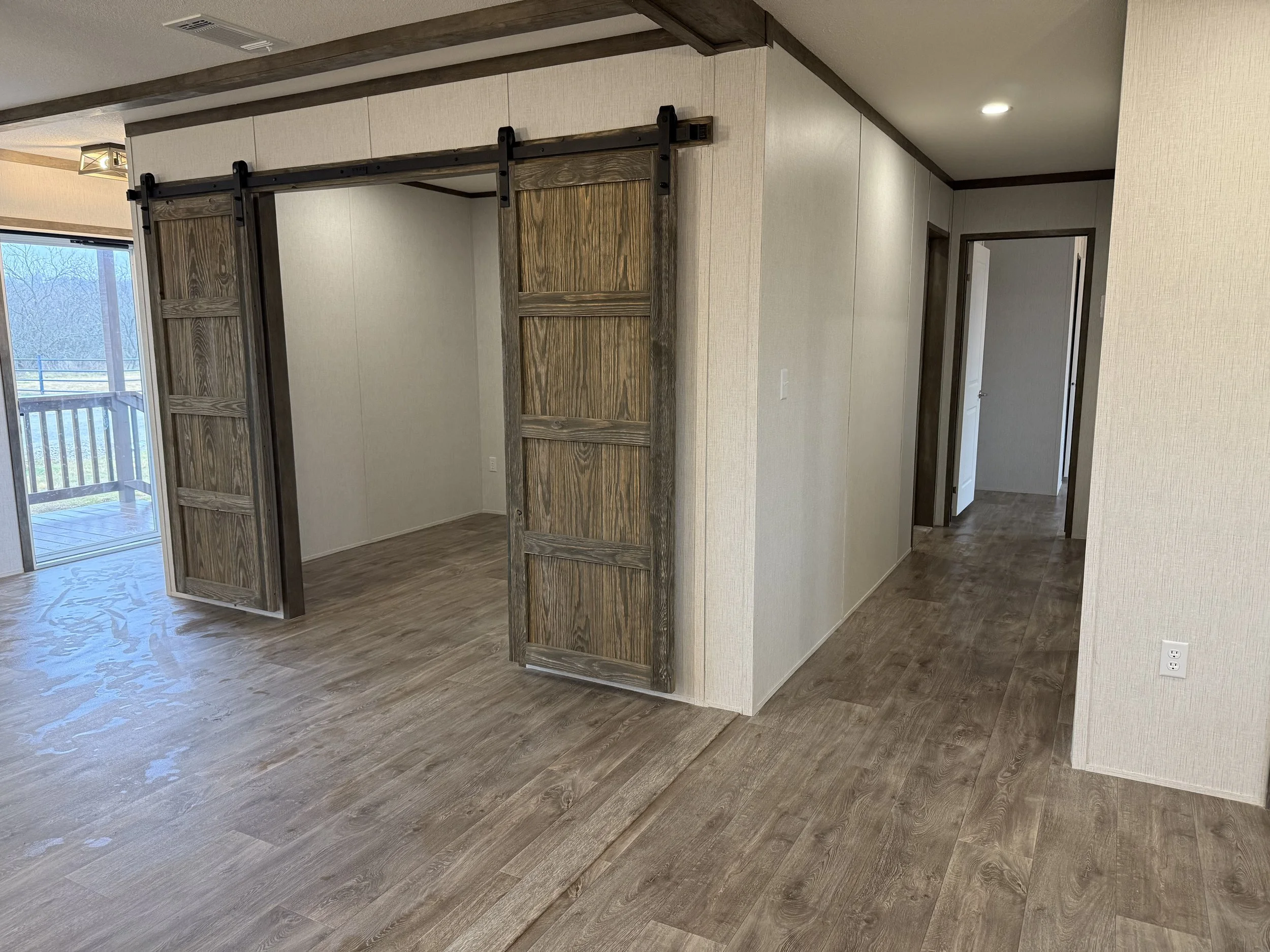 Interior view of a room with wood flooring, a small closet with sliding barn doors, and a doorway leading to a hallway. A sliding glass door to a balcony is visible on the left side.