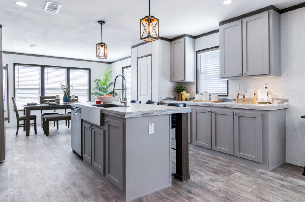 Modern kitchen with light gray cabinets, marble countertops, and a kitchen island with a built-in sink. There are pendant lights hanging from the ceiling, and a dining area with a table and chairs near windows with blinds. A plant is on the counter, 