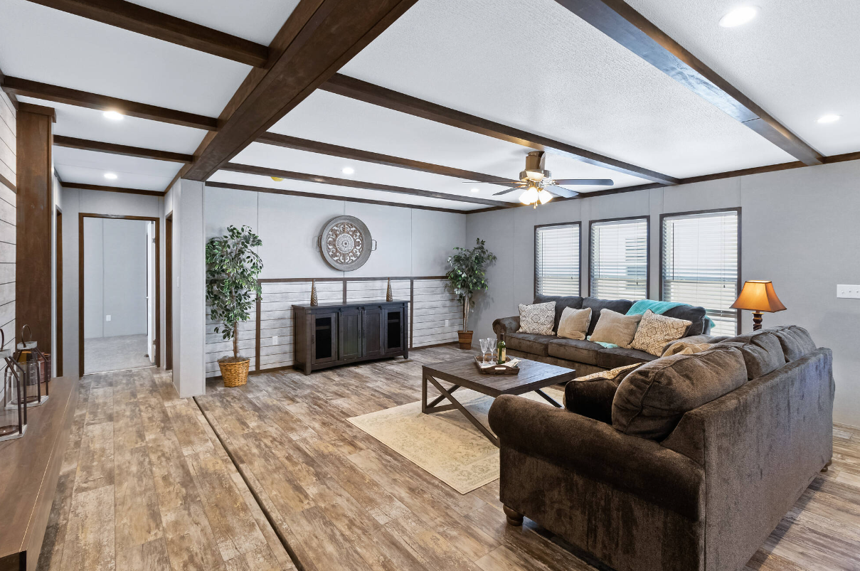 Living room with wood flooring, two brown sofas, a wooden coffee table, and multiple windows with blinds. There are potted plants and a ceiling fan with lights.