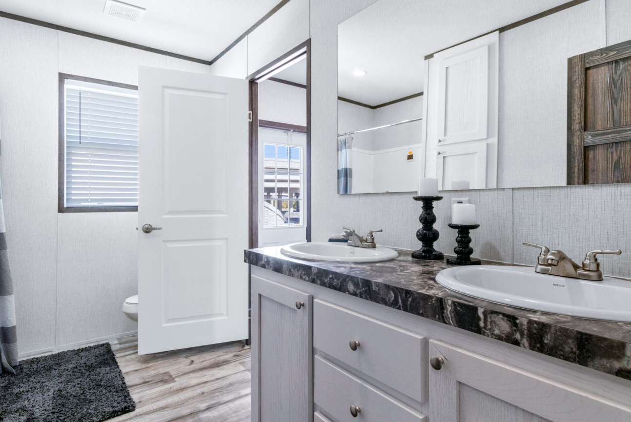 A bathroom with a double sink vanity, candles on decorative holders, a mirror, and a window with blinds, with a door leading to a shower or bathtub area.