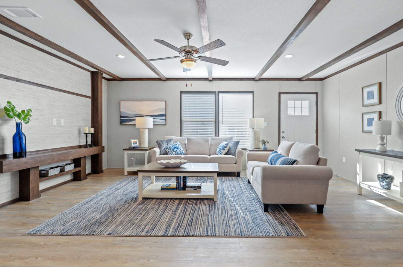 Bright living room with two beige sofas, a wooden coffee table with books, a blue area rug, and various white and blue decorative items and lamps. There are two windows with blinds, a ceiling fan, and framed artwork on the walls.