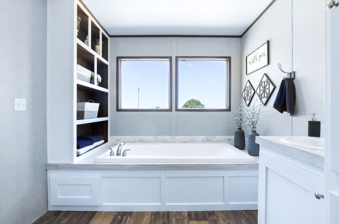 Bathroom with a built-in bathtub, two windows, open shelving with baskets, decorative wall art, and vases with flowers.