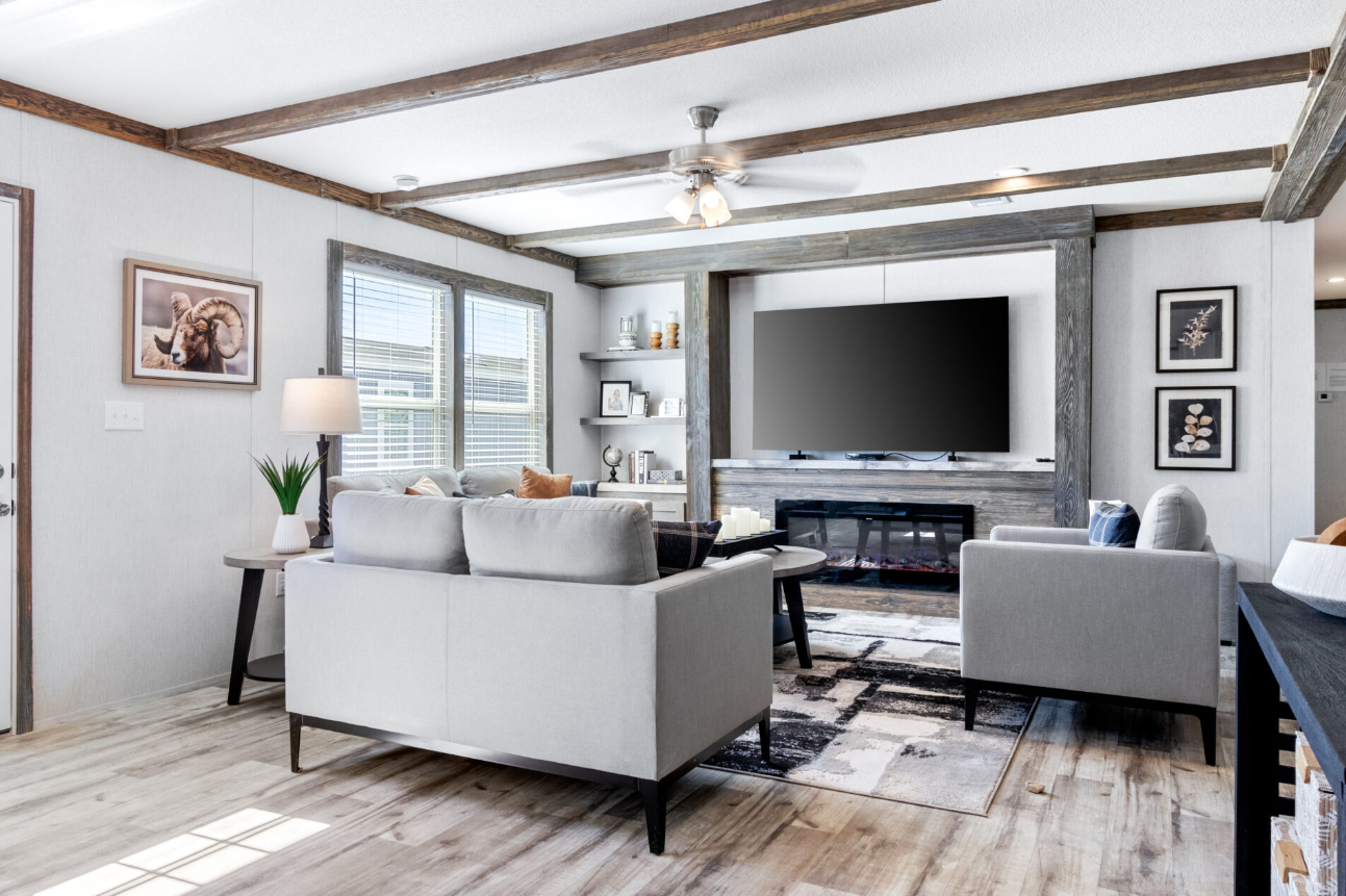 Modern living room with white sofas, a wall-mounted flat-screen TV, wooden ceiling beams, and natural light from windows.