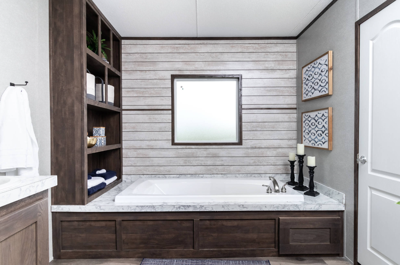Bathroom with a built-in bathtub, wooden shelves with towels and decor, candle holders, framed blue patterned artwork, and a frosted window.