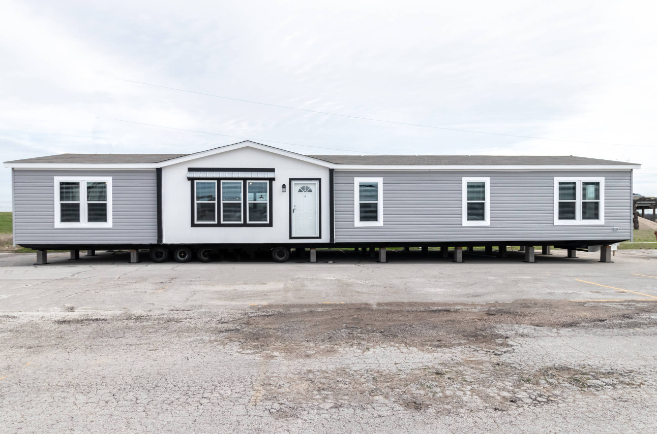 A gray manufactured home with white trim, elevated on supports, located in a parking lot under a cloudy sky.