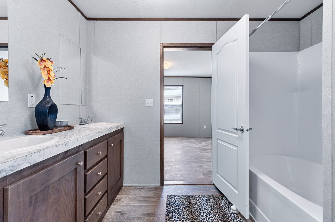 Bathroom with vanity and double sink, vase with flowers, mirror, and a shower stall with curtain, leading to a room with a window and wood flooring.
