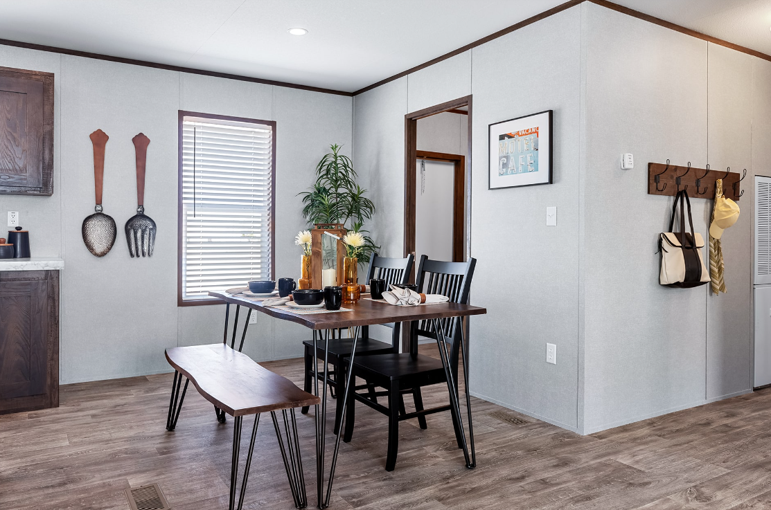 Dining area with a wooden table, black chairs, a bench, and decorative vases, with a window and wall decor including large utensils, a framed sign, and a hook rack with bags and hats.