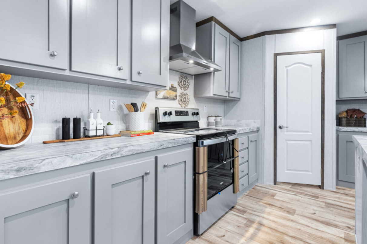 Light gray kitchen with marble countertops, gray cabinets, stainless steel range hood, and a stove with ties on the oven handle. Decor includes a round tray with flowers, black salt and pepper shakers, white bottles, and a cutting board, with wooden 