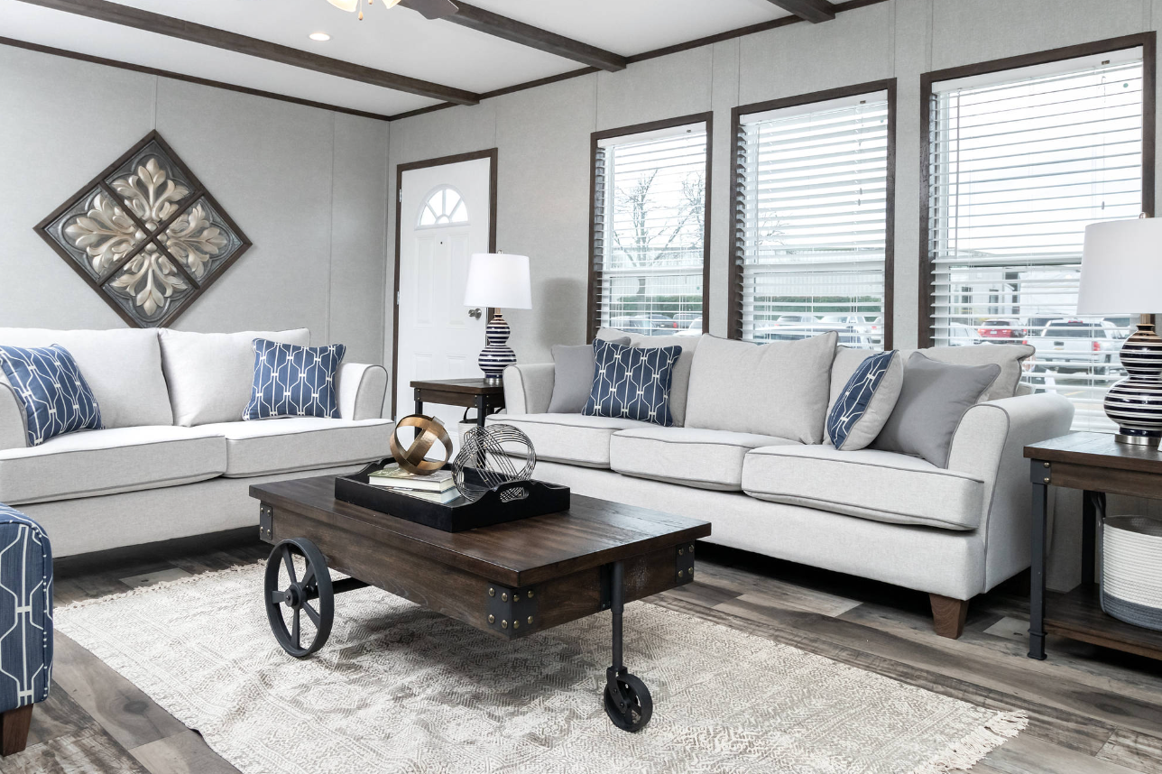 Living room with two white sofas with blue patterned pillows, a wooden coffee table with decorative objects, two side tables with lamps, and windows with blinds.