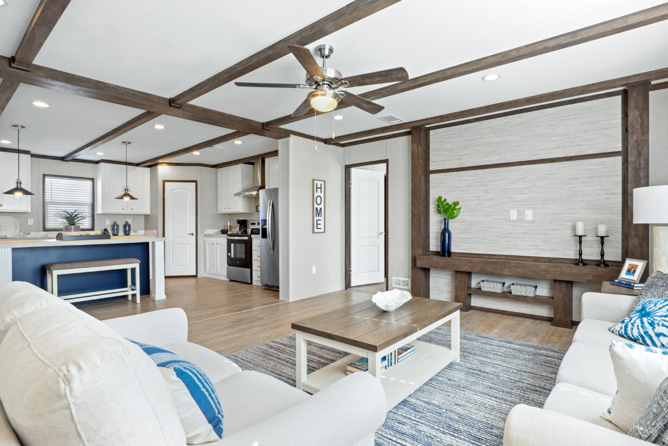 Open concept living room with white sofas, wooden coffee table, and a partial view of the kitchen with white cabinets and stainless steel appliances, including a refrigerator and oven. The ceiling features exposed wooden beams and a ceiling fan. Deco