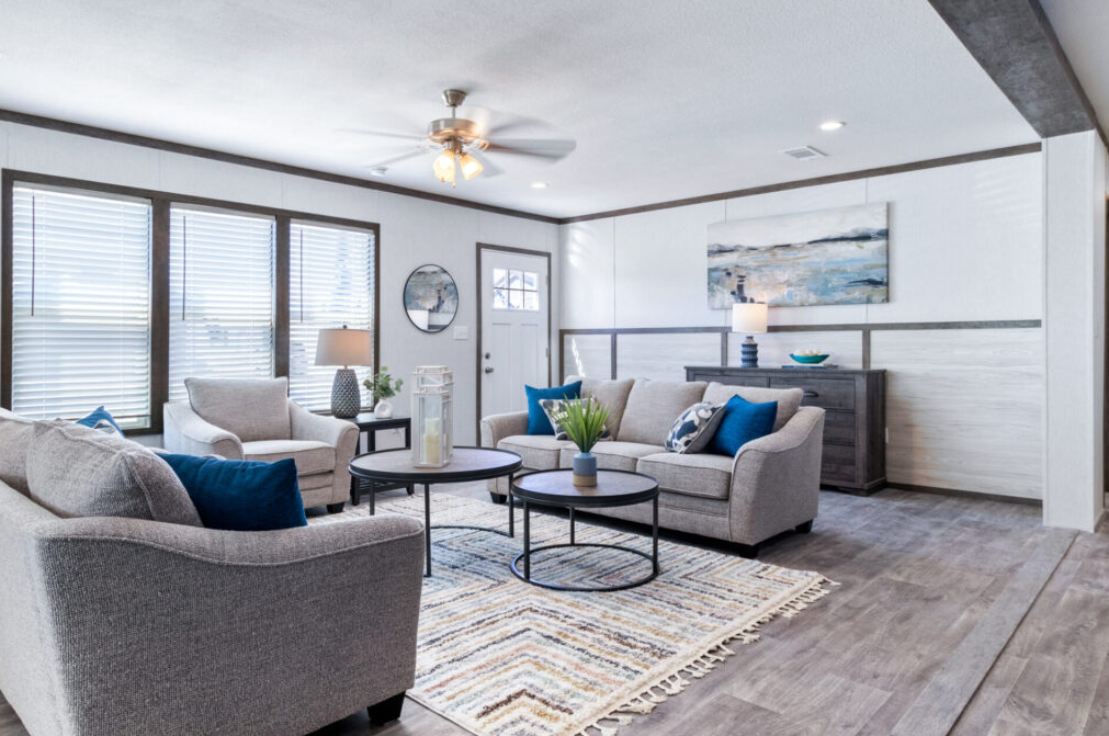 Living room with three light-colored sofas, blue pillows, a striped area rug, large windows with blinds, a ceiling fan, and a sideboard with a lamp and artwork.