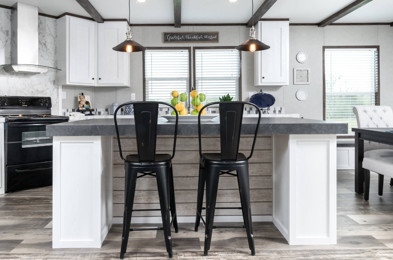 Modern kitchen with black and white decor, island with two black chairs, fruit centerpiece, and windows with blinds