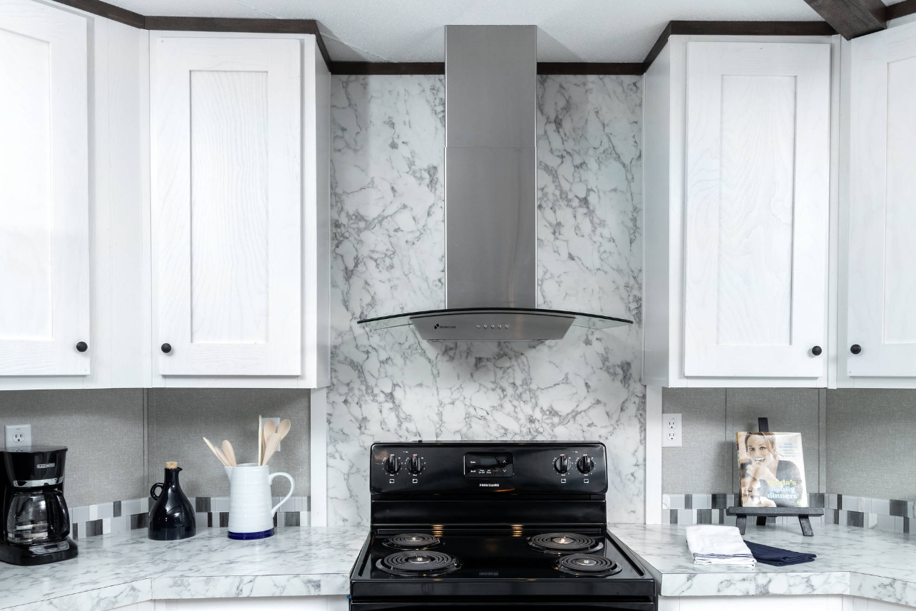 Kitchen with white cabinets, marble backsplash and countertops, a black electric stove with four burners, a stainless steel range hood, a coffee maker, a black vase, a white utensil holder with wooden utensils, a magazine on a stand, and a stack of w