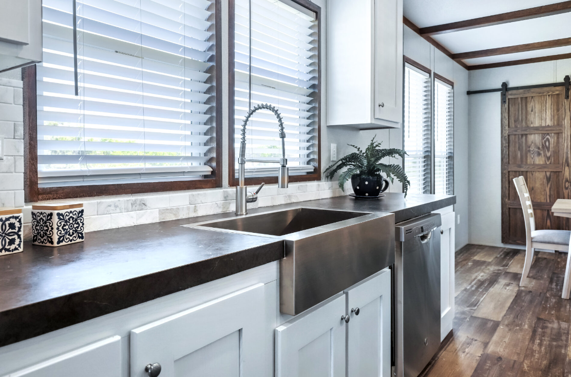 Kitchen with white cabinets, a black countertop, stainless steel farmhouse sink, wooden-framed windows with blinds, and a potted plant on the counter.