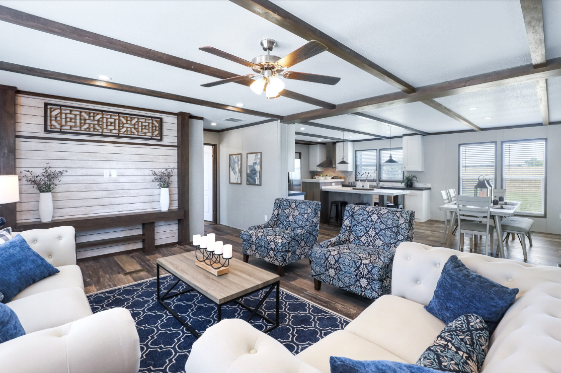 Open-concept living room and kitchen with white and blue decor, wood accents, ceiling fan, and large windows letting in natural light.