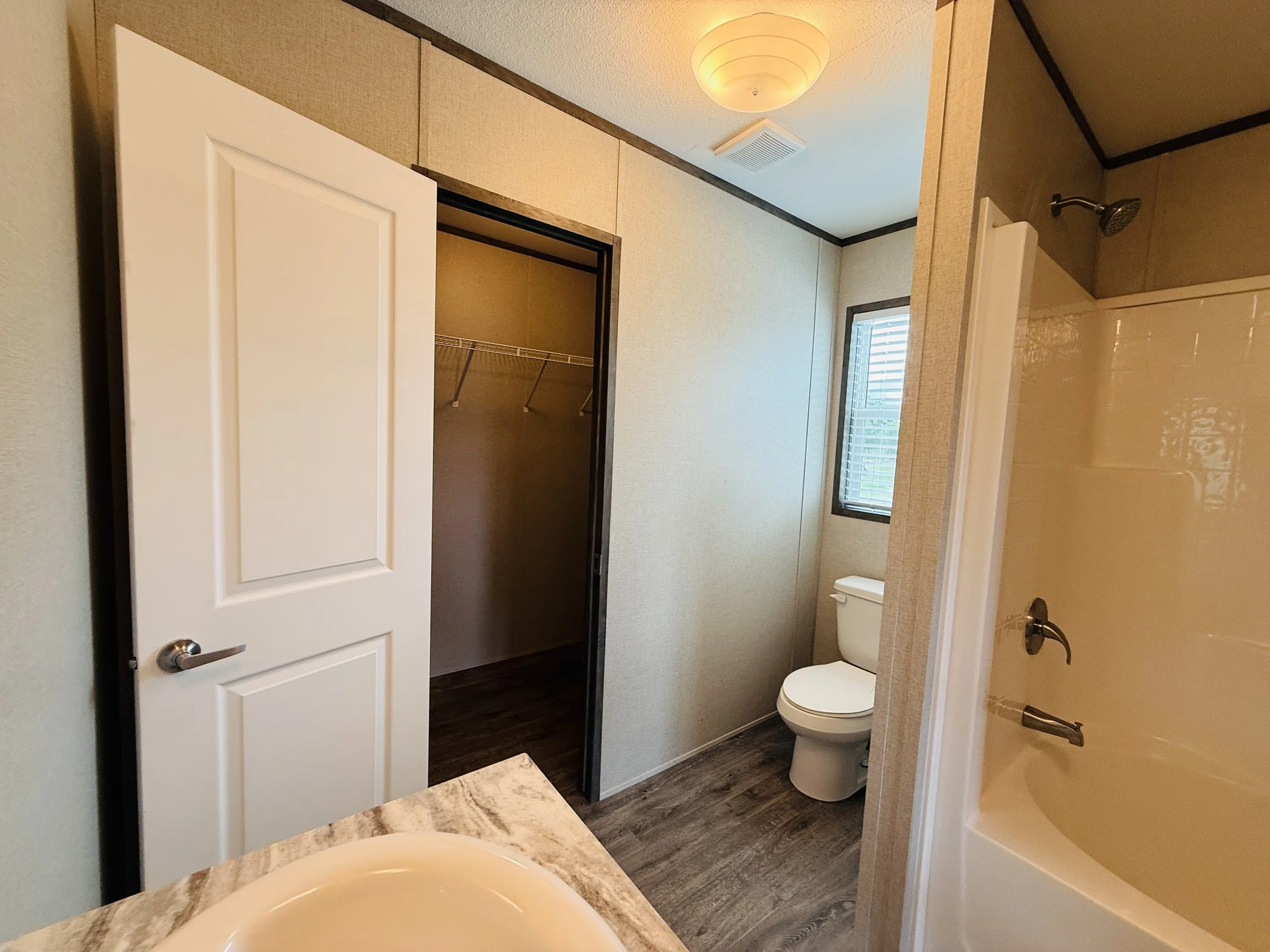 Small bathroom with beige walls, a window, a white toilet, a bathtub with a shower, a marble countertop with a sink, and a closet with an open door showing a wire shelf inside.