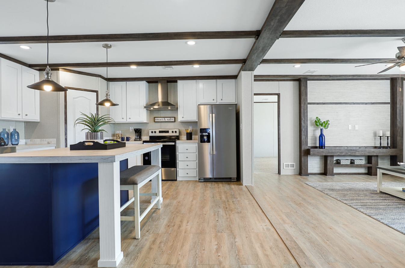 Open-concept kitchen and living area with white cabinets, a stainless steel refrigerator, a navy blue kitchen island, and wooden beams on the ceiling.