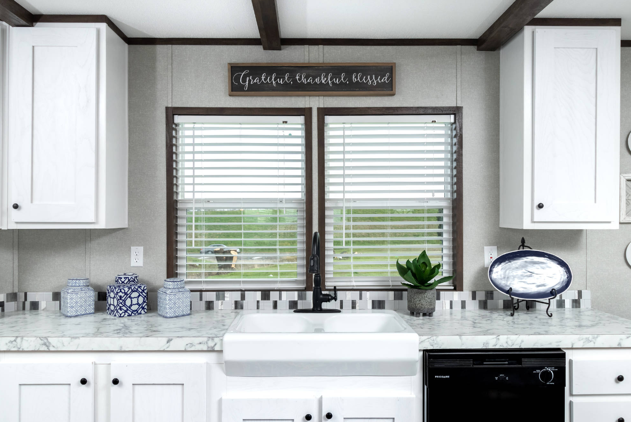 Kitchen with two windows, white cabinets, marble countertop, black faucet, decorative jars, a potted plant, a dish on a stand, and a sign that reads 'Grateful, thankful, blessed' above the windows.