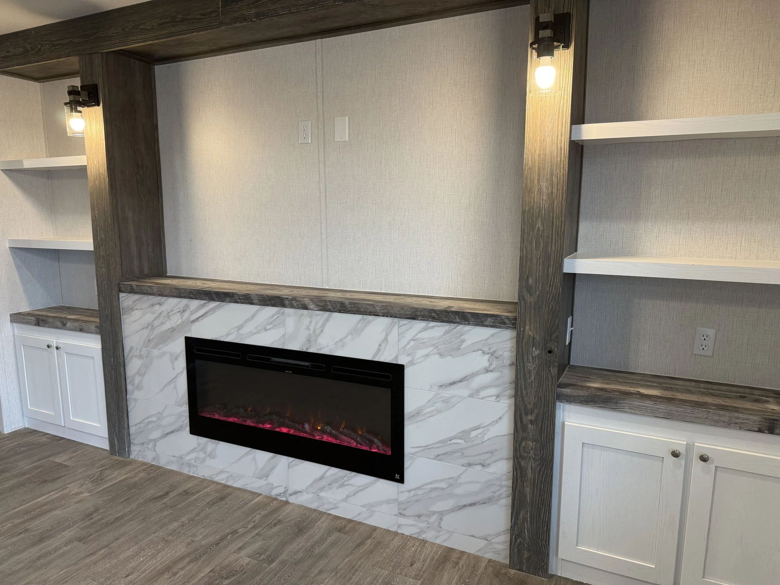 Living room wall with built-in electric fireplace surrounded by marble tile, wooden shelves on each side, and cabinets below. Wall sconces with exposed bulbs above the shelves.
