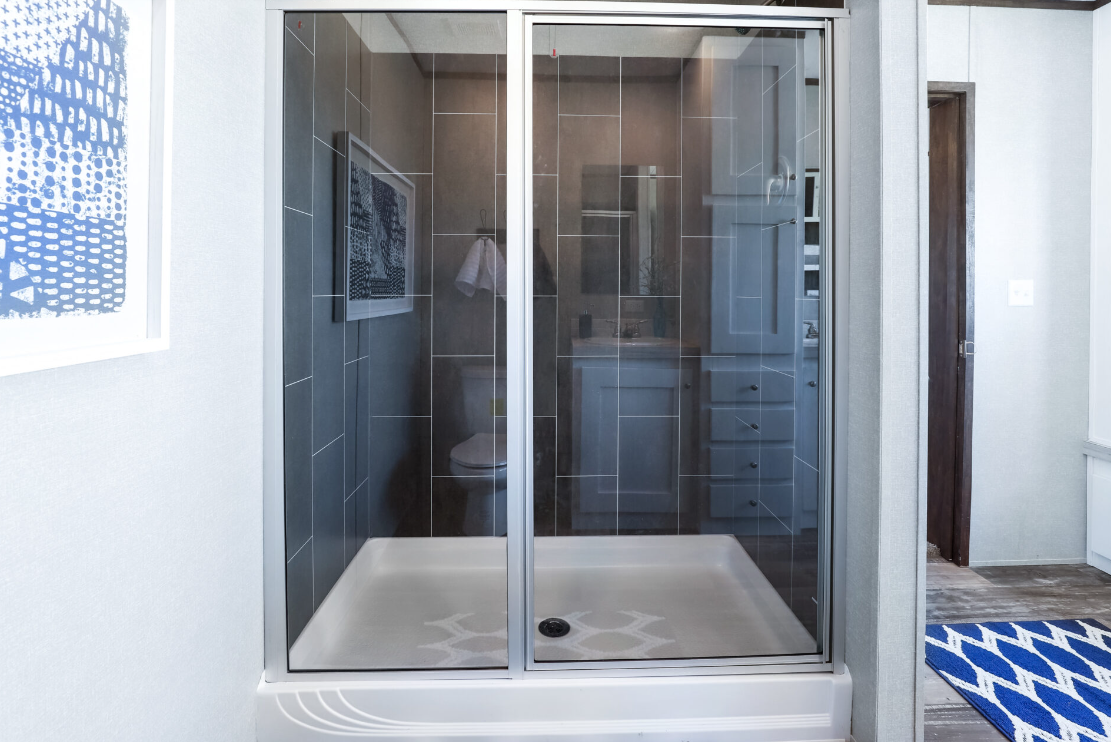 A modern glass shower with gray tiled walls, a white shower base, and a white towel hanging inside. The shower is in a bathroom with light-colored walls, a blue and white patterned rug, and a piece of abstract art on the wall.