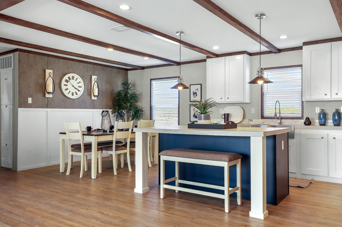 Open-concept kitchen and dining area with white cabinetry, a kitchen island with a blue base, and a wooden dining table with six chairs. Decorative items, wall clock, and houseplants are visible.