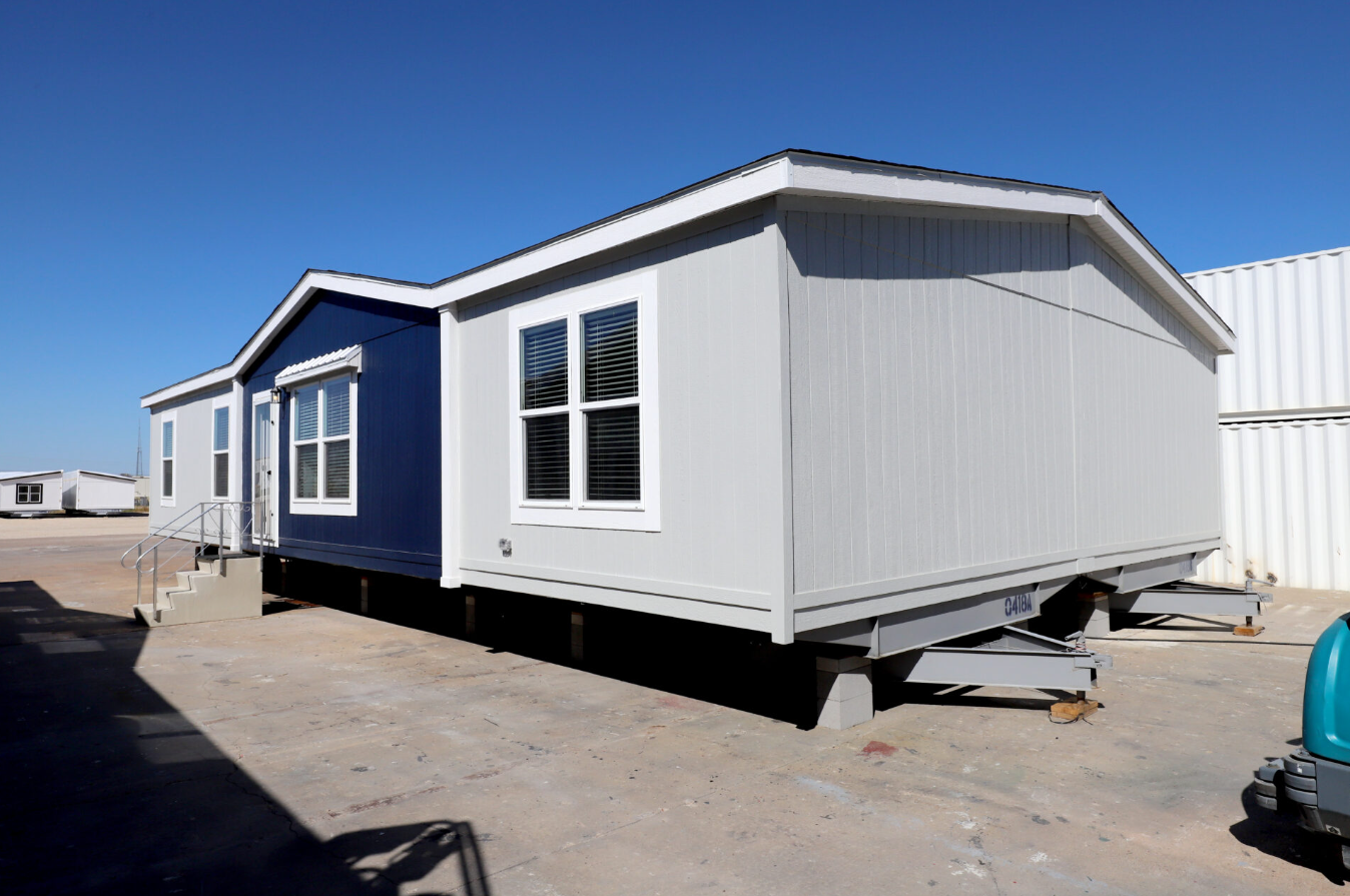 A manufactured mobile home with white walls and dark blue accents, elevated on concrete blocks on a gravel lot under a clear blue sky.
